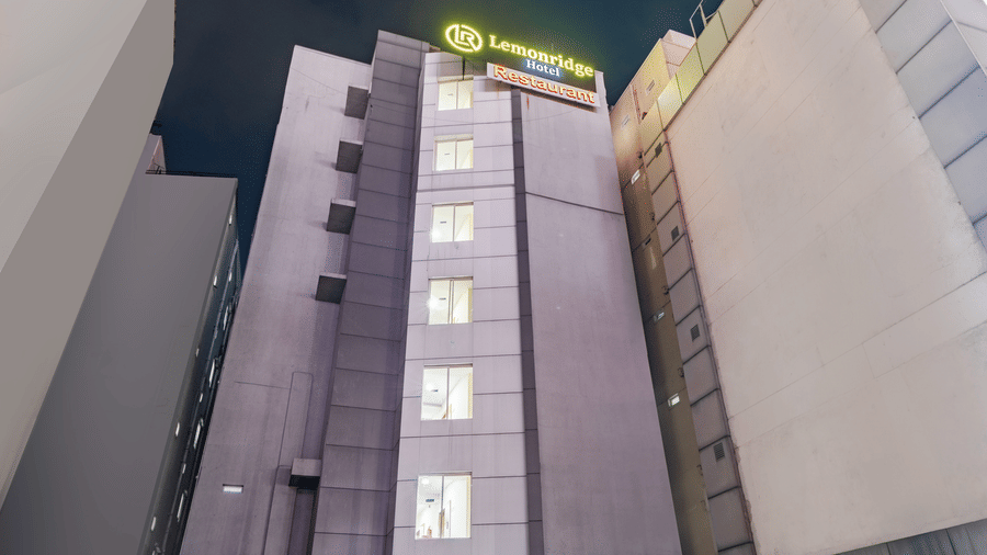 A low angle view at night shows the tall, multi storey concrete exterior structure and illuminated roof signage of Lemonridge by Monday Hotels - Kukatpally.