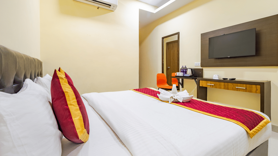 A large bed with decorative cushions faces a wall mounted television, a wooden desk, and an air conditioning unit at Lemonridge by Monday Hotels - Kukatpally.
