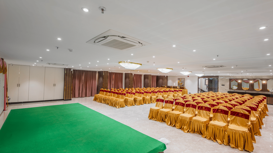 A spacious banquet hall at Lemonridge by Monday Hotels - KPHB featuring a green carpeted front stage, rows of chairs wrapped in gold covers, decorative curtains, and ceiling spotlights.