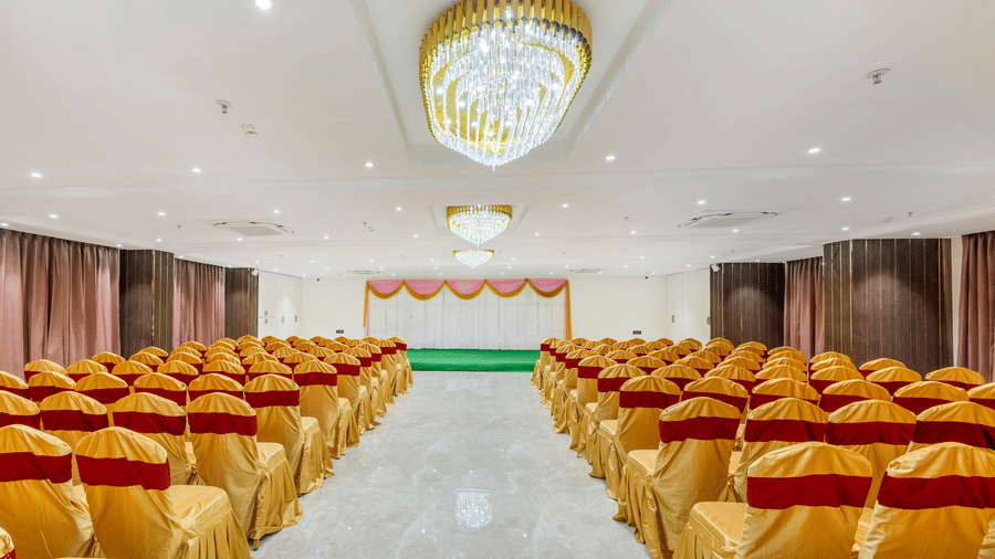 Banquet hall at Lemonridge by Monday Hotels - KPHB featuring hanging crystal chandeliers,rows of chairs, and a small stage.