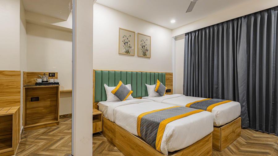 Lemonridge Hotels, KPHB |A bedroom at Lemonridge by Monday Hotels - KPHB featuring twin bed, green headboard, wall pictures, dressing mirror, and luggage table. Hotel room with two beds, side table, and a window with curtains. Simple and functional.