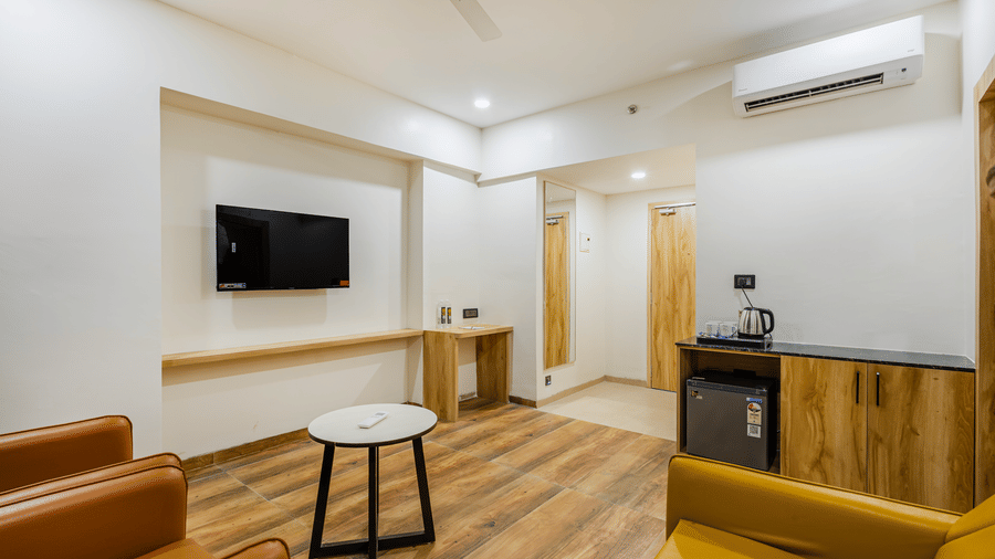 The living room of a suite at Lemonridge by Monday Hotels - KPHB, featuring an armchair, a yellow sofa, a coffee table, a TV, and a mini-fridge counter.