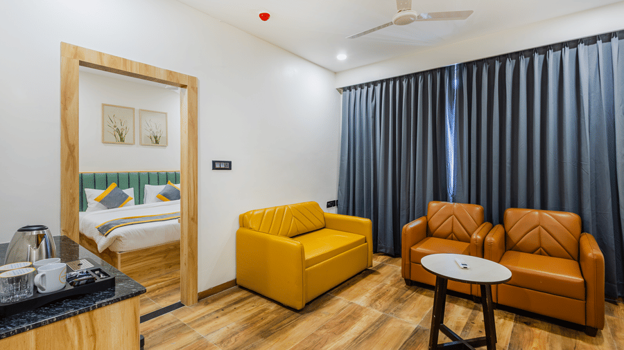 A room at Lemonridge by Monday Hotels - KPHB featuring a separate living area with a  yellow sofa, armchairs, round table, tea station, vanity mirror, and bedroom visible through the doorway.