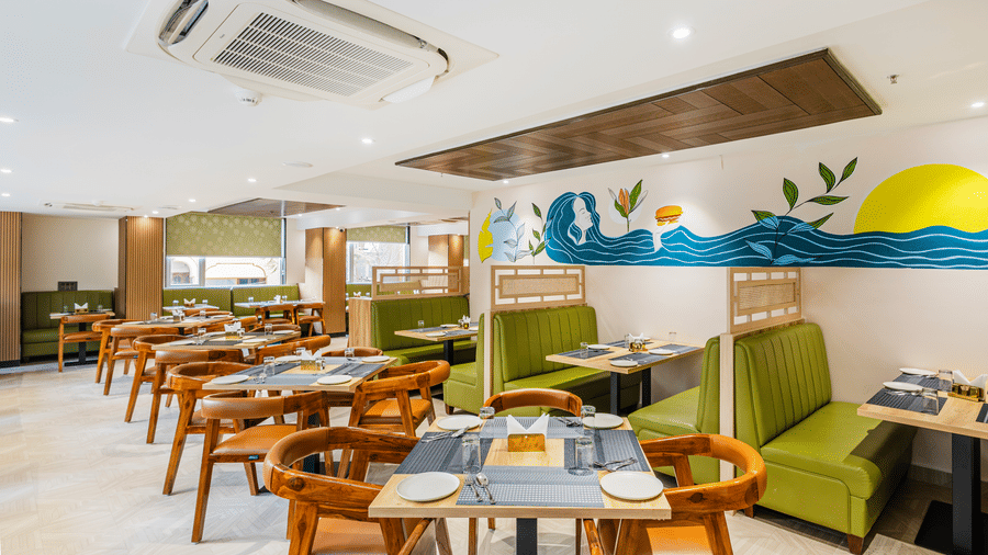 Curry Leaf restaurant at Lemonridge by Monday Hotels - KPHB featuring green booth seating, wooden tables, chairs, and a colorful wave wall mural.