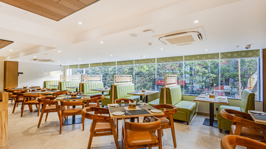 Curry Leaf restaurant at Lemonridge by Monday Hotels - KPHB featuring wooden tables, cutlery sets, green booth seating, and wide windows.