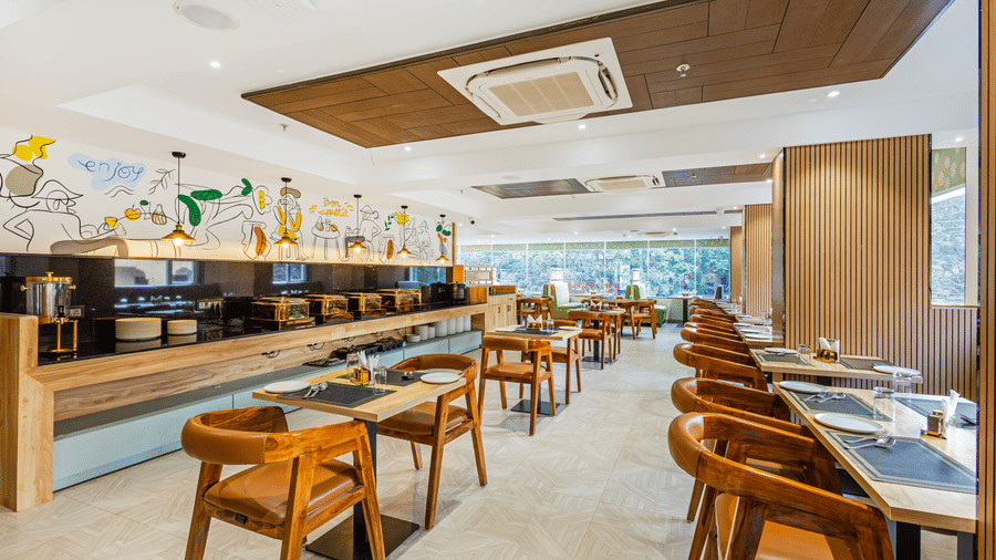 Curry Leaf restaurant at Lemonridge by Monday Hotels - KPHB featuring a long buffet station, wooden tables, chairs, and a ceiling AC unit.