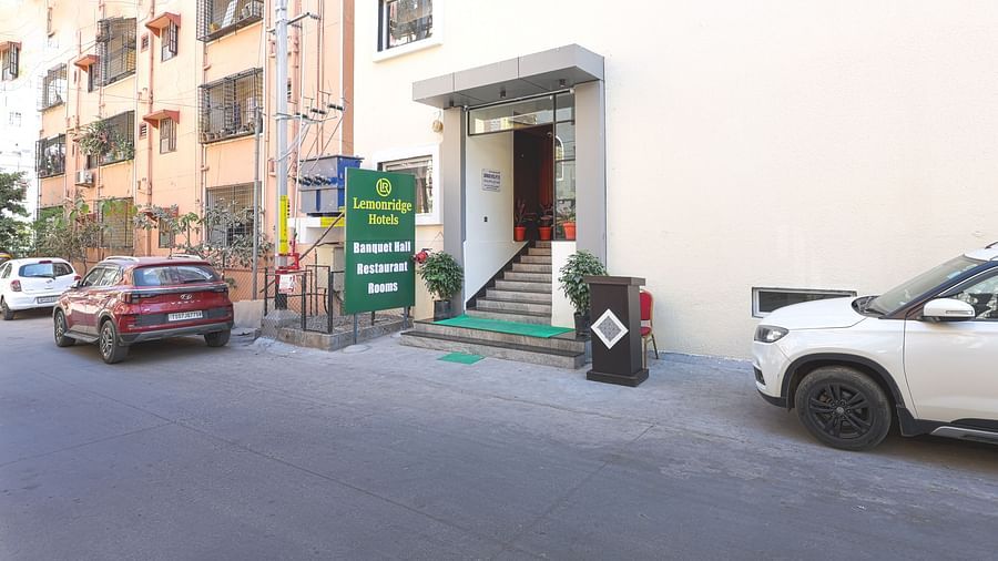 The entrance to the Lemonridge Hotels KPHB property, featuring a signboard that reads 'Lemonridge Hotels' at the top and 'Banquet Halls, Restaurant, Rooms' below. Other buildings are visible on the side, with vehicles parked outside.
