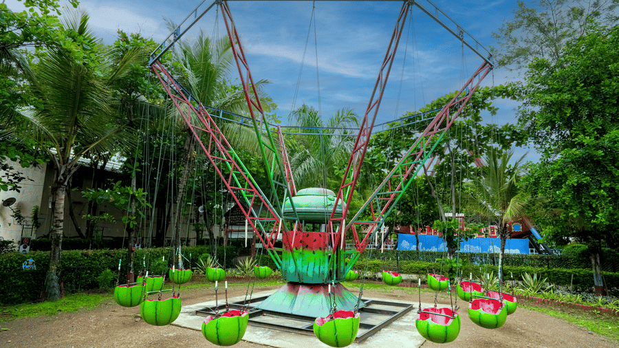 A colourful outdoor swing carousel with suspended seats, set within a landscaped garden surrounded by trees.