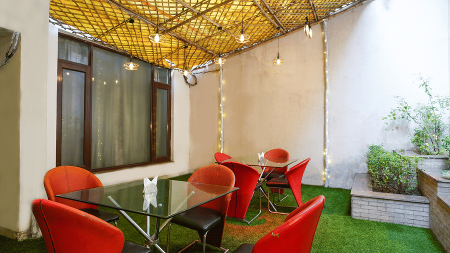 Outdoor courtyard cafe with red tub chairs, glass-top tables, a bamboo-lattice canopy strung with fairy lights, and grass.