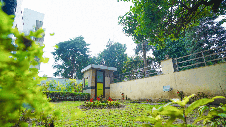 Garden with a lawn, a red flower bed, a LimeTree-branded guard cabin, a boundary wall with railing, and trees against the sky.