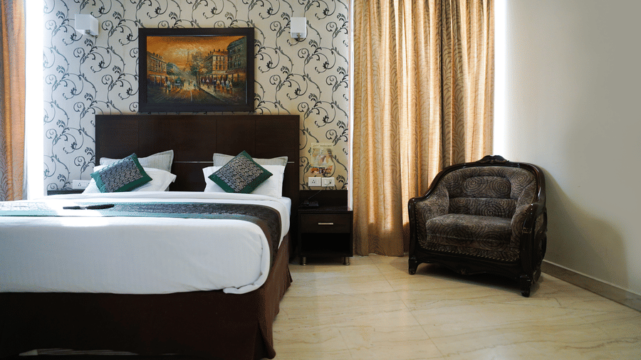 Guest room with a dark wood headboard, a framed Parisian painting, damask wallpaper, gold curtains, and a carved armchair.