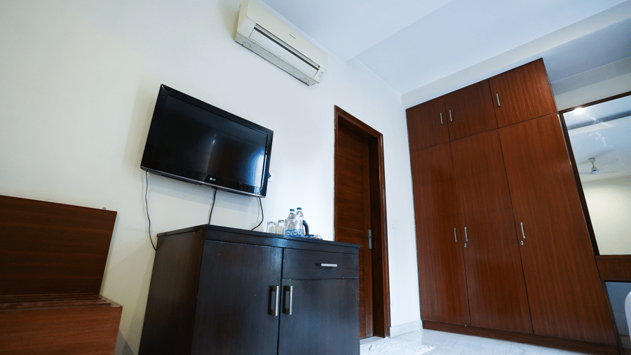 Guest room detail with a wall-mounted LG TV, a split air conditioner, a dark minibar with water bottles, and a wood wardrobe.