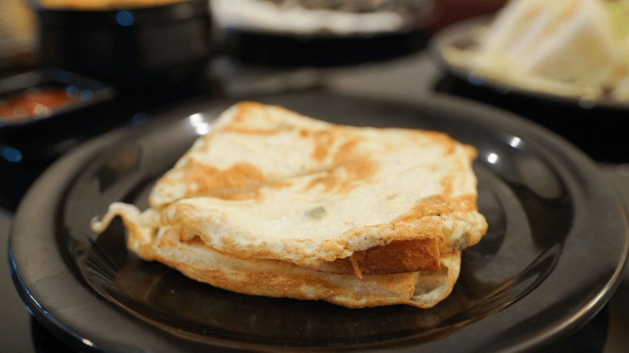 A toasted sandwich served on a black plate, with other plates of food blurred in the background on a dining table.