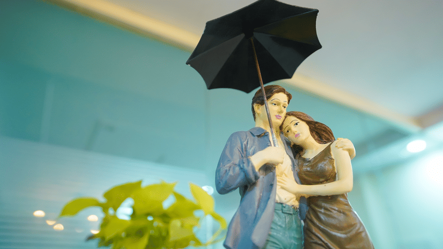 Decorative figurine of a couple standing close together under a black umbrella, displayed near a plant on a glass surface indoors