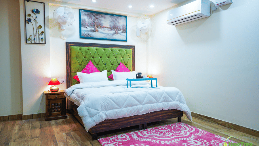 Guest room with a green tufted headboard, framed snowy landscape, wall fans, red lamp, and a pink patterned rug at Lime Tree Hotel Near 32nd Avenue.
