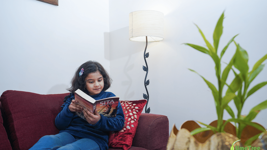 A child in blue reads the book SITA on a maroon sofa beside a floor lamp and gold planter at Lime Tree Hotel Near 32nd Avenue's seating corner.