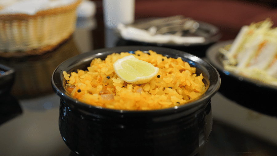 Bowl of yellow lemon rice garnished with a lemon wedge and mustard seeds, with a bread basket and sandwiches in the background