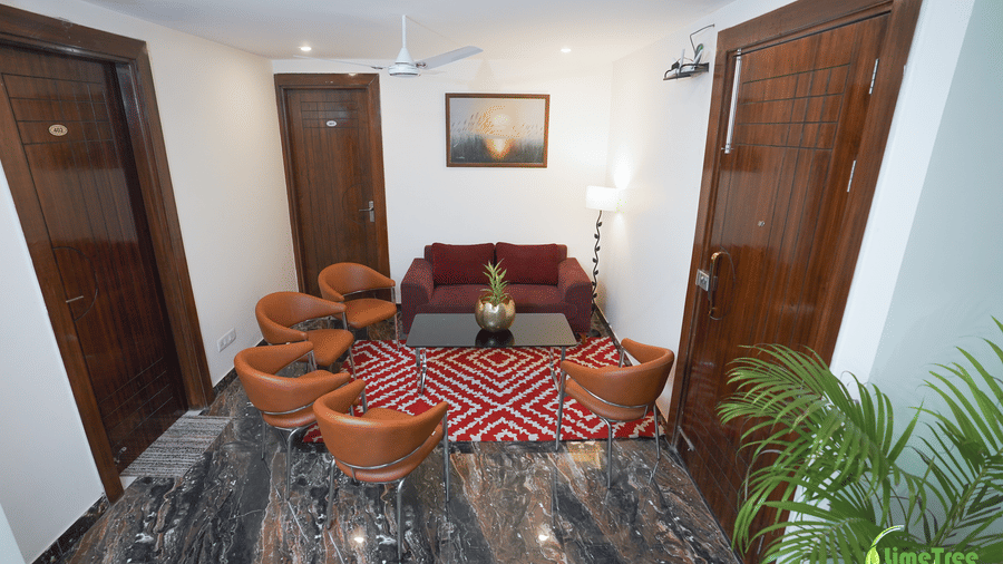 Lounge seating at Lime Tree Hotel Near 32nd Avenue with a maroon sofa, 4 tan leather chairs, a glass table, and a red rug on marble flooring.