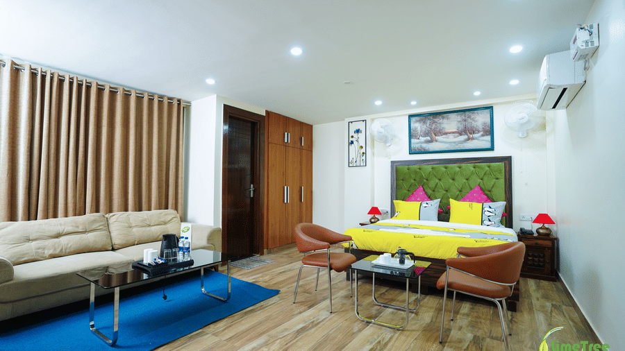 A spacious hotel room layout showing a sofa seating area on the left and a double bed with yellow linen on the right.