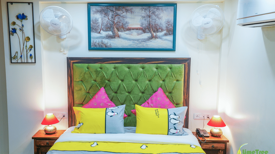 A hotel bedroom featuring a double bed with colourful yellow and pink cushions, a green tufted headboard, and two bedside tables.