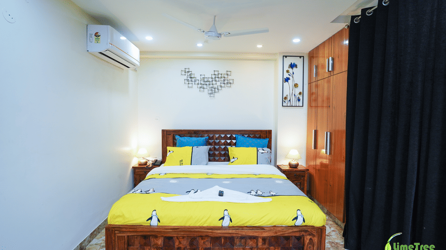 Bedroom at Lime Tree Hotel Near 32nd Avenue with a wooden bed, penguin-print bedding, blue cushions, metal heart wall art, and a split AC.
