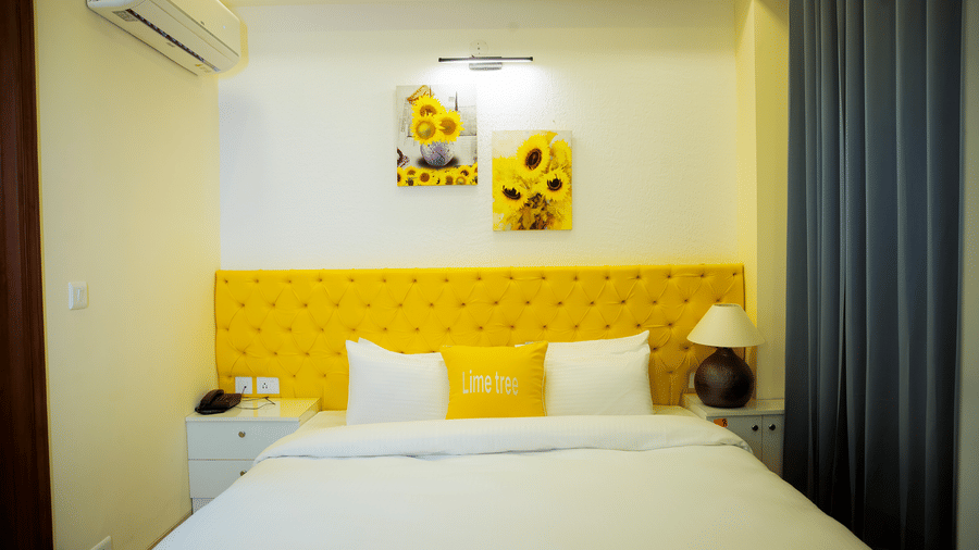 Bedroom of Lime Tree Hotels & Service Apartment Private Limited featuring a yellow headboard, white bedding, and dark curtains.
