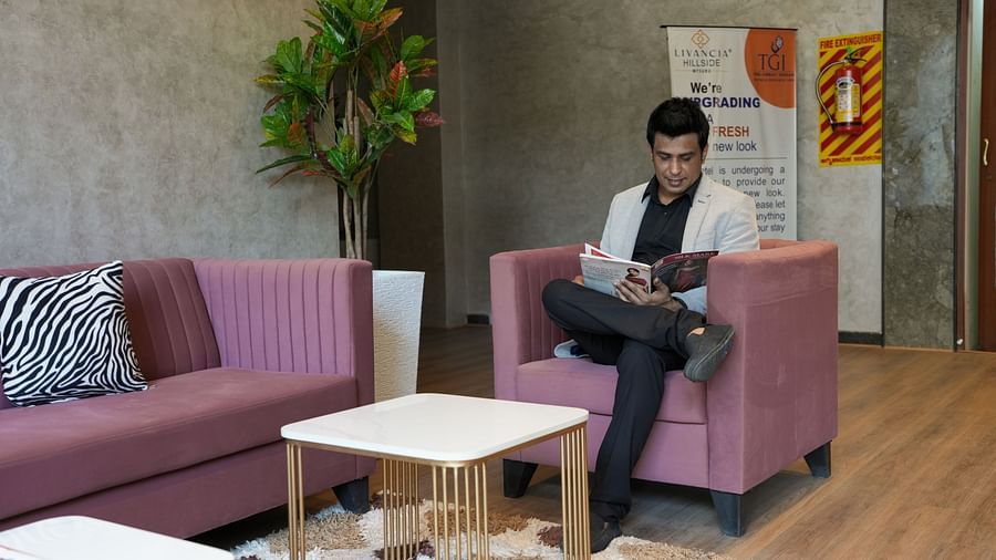 Medium shot of a man seated in the lounge area reading a magazine, surrounded by pink seating and indoor plants at TGI Livancia Hillside, JC Nagar, Mysore
