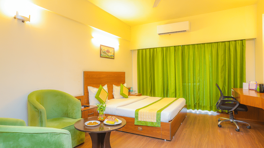 Wide view of the Premium Room with Balcony at Livbox, Dehradun, furnished with a king size bed, a seating area with 2 upholstered chairs, ample lighting, a work desk, and closed curtains.