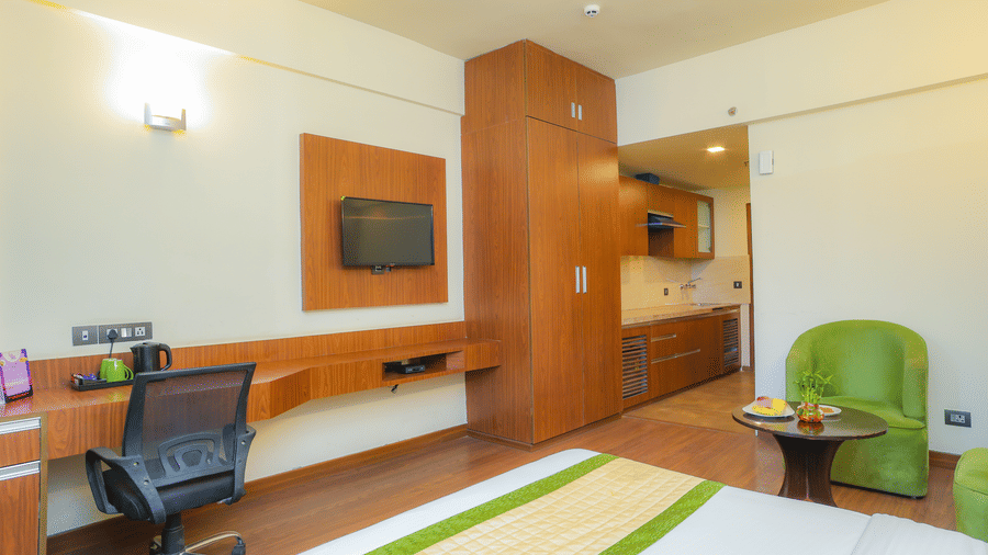 Corner view of the Premium Room with Balcony that is furnished with a king size bed, a seating area, a wall-mounted TV, a wardrobe, a kitchenette, and wooden flooring.