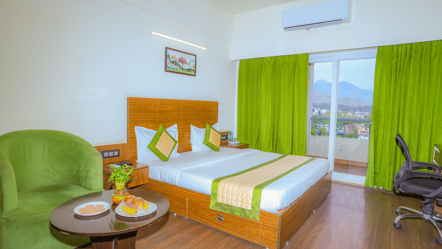 A king size bed in the Premium Room with Balcony at Livbox, Dehradun, which also features wooden flooring, curtained windows opening to the views of the surrounding, and a seating area.