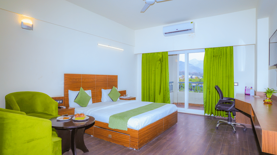King size bed, wooden flooring, drawn curtains, seating area with upholstered chairs, and a work desk in the Suite With Balcony at Livbox, Dehradun.