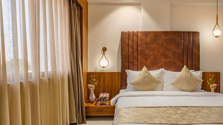 A comfortable bed is situated between two nightstands with lamps in a hotel room with layered window coverings - Lords Eco Inn Ambaji.