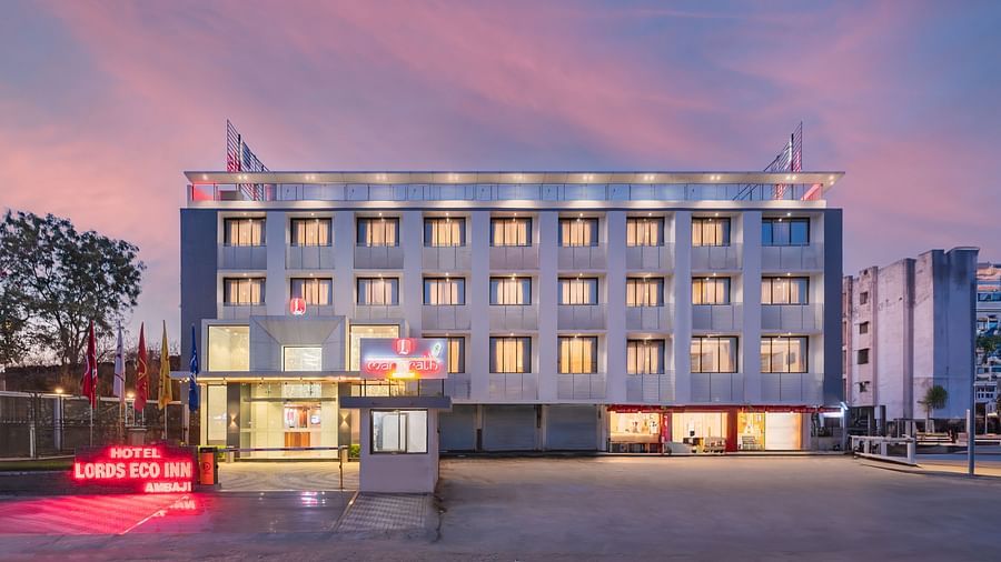 A modern hotel building with many windows and a bright red sign stands beside a paved area under a twilight sky - Lords Eco Inn Ambaji.