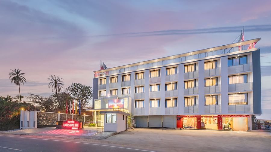 A multi-story building with illuminated windows and a red sign stands beside a paved area at dusk - Lords Eco Inn Ambaji.