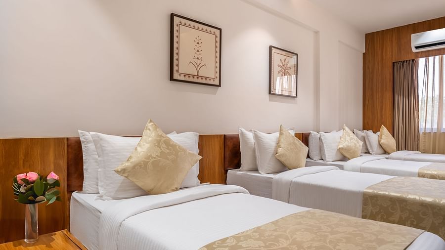 Three single beds with white linens and decorative pillows line a hotel room with framed artwork and a window - Lords Eco Inn Ambaji.