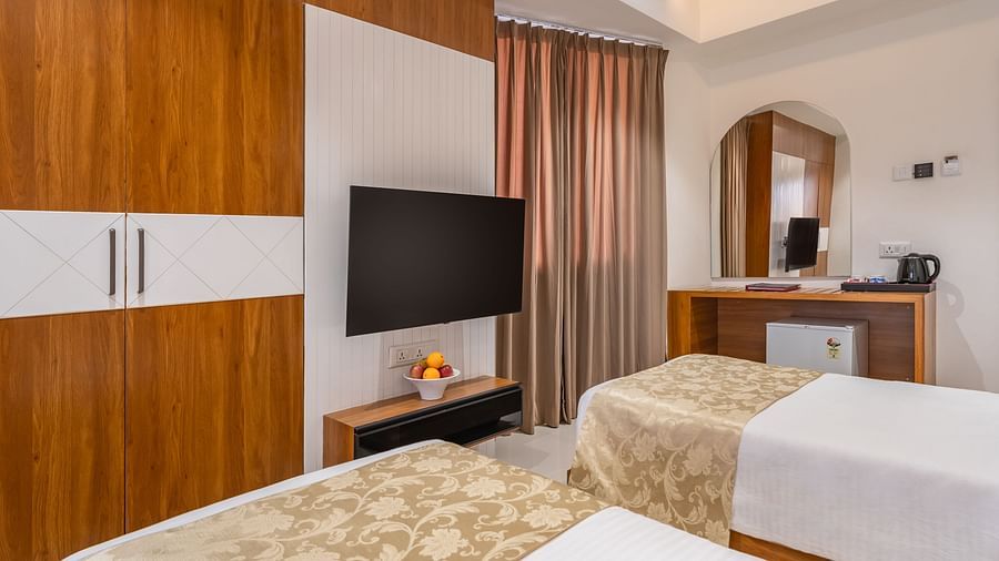 A hotel room features two single beds, a wall-mounted television, a wardrobe, and a desk with a mirror - Lords Eco Inn Ambaji.