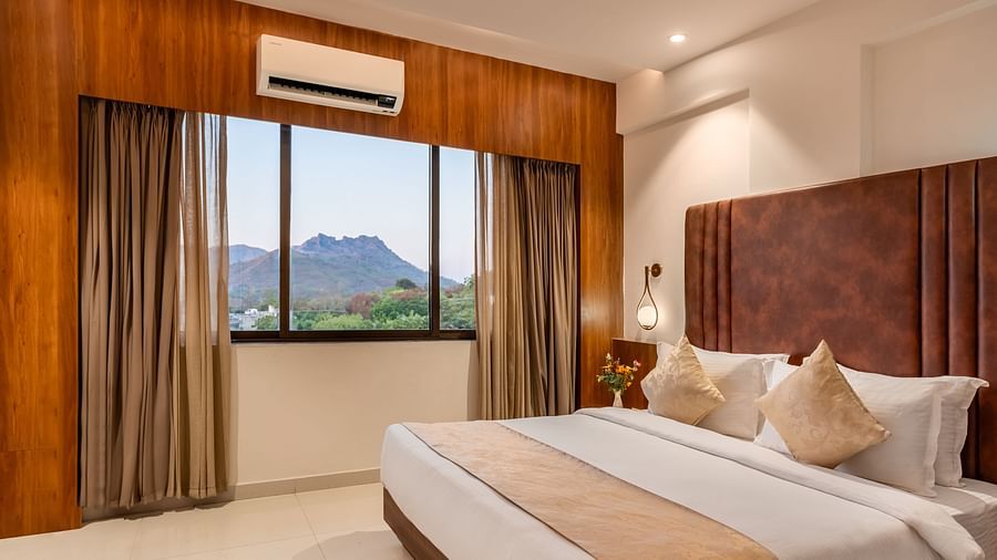 A comfortable king-size bed with soft pillows and a throw faces a window with curtains revealing a mountain view - Lords Eco Inn Ambaji.