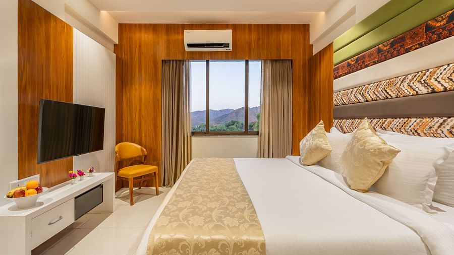 A well-appointed hotel room features a king-size bed, a mounted television, a cushioned chair, and a view through the window - Lords Eco Inn Ambaji.