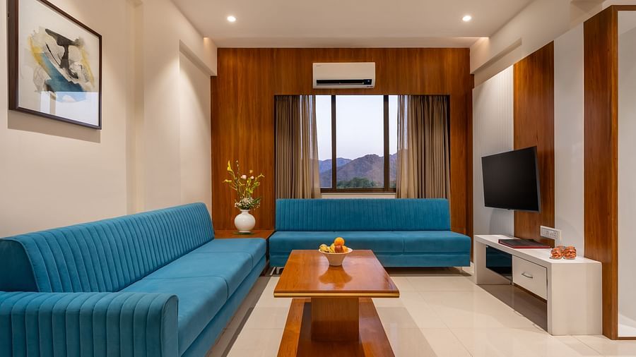 A living area features two sofas, a wooden coffee table with fruit, a television, and framed artwork - Lords Eco Inn Ambaji.