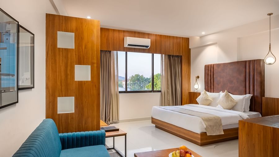 A well-lit hotel suite features a comfortable bed, a sofa, a wooden coffee table with fruit, and a window with curtains - Lords Eco Inn Ambaji.