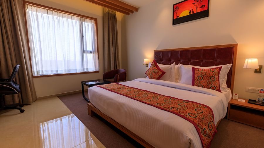A well-lit hotel room at Lords Eco Inn Apple Gate features a double bed with patterned accents, a desk, a chair, and window curtains.