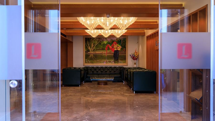 Through glass doors marked with a red L, a warmly lit hotel lobby with seating, artwork, and a distinctive chandelier is visible at Lords Eco Inn Apple Gate