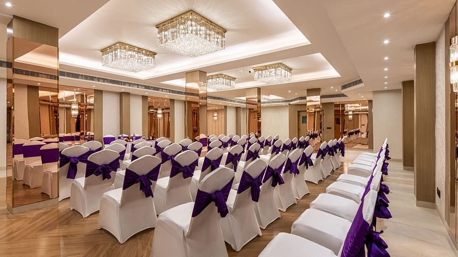 An event space with rows of white chairs decorated with purple fabric, ready for guests. It features a decorative ceiling with hanging lights and mirrored wall accents - Lords Eco Inn Dehradun.