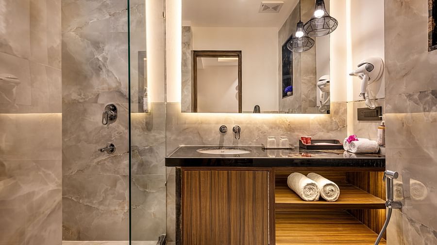 A modern bathroom with a glass-enclosed shower, dark countertop, and a large mirror with lighting. It features a wooden cabinet with open shelves, hanging pendant lights, and marble-like tile walls - Lords Eco Inn Dehradun.
