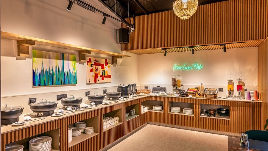 A buffet area with serving dishes, offering a variety of food and drinks, and a hanging chandelier. It features wooden paneling, colourful artwork on the wall, and a neon sign - Lords Eco Inn Dehradun.