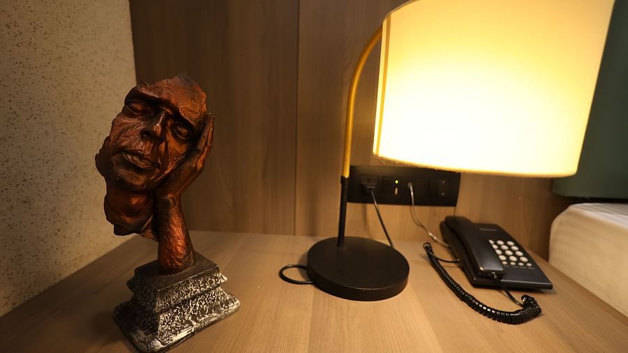 The image shows a bedside table with a lamp illuminating a sculpture of a face and a telephone - Lords Eco Inn Navsari.