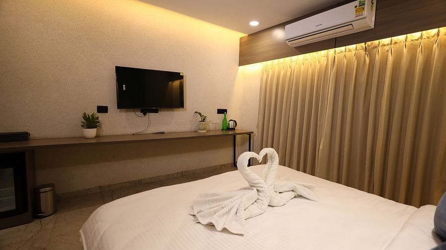 The image shows a hotel room with a bed featuring swan-shaped towel art, a television, a desk, and curtains - Lords Eco Inn Navsari.