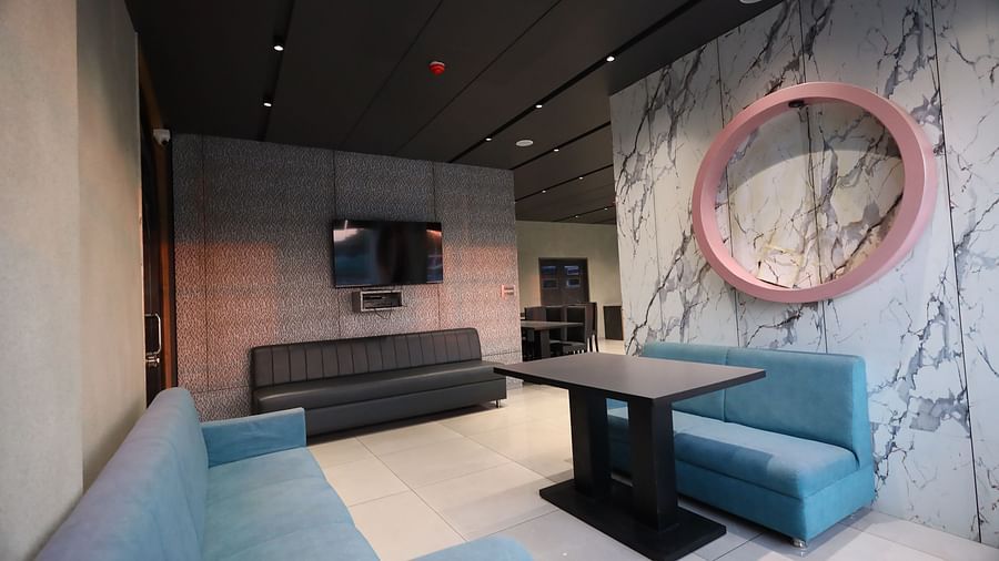 The image shows a modern lounge area with sofas, a table, a television, and a circular mirror on a patterned wall - Lords Eco Inn Navsari.