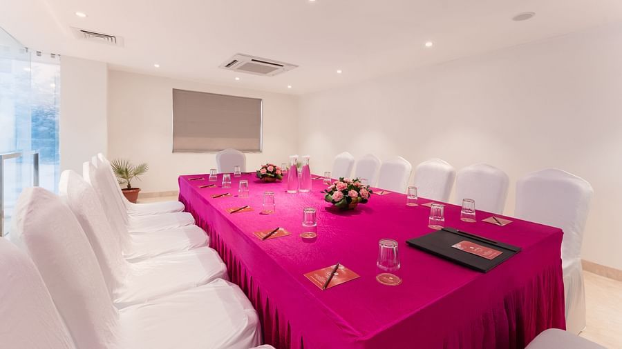 A well-lit meeting room at Lords Inn Udaipur is set with a vibrant pink tablecloth, white chairs, water glasses, and floral centerpieces.