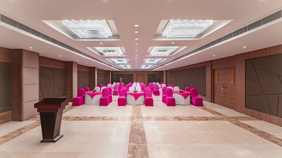 A spacious and well-lit banquet hall at Lords Inn Udaipur is arranged with rows of chairs covered in vibrant pink fabric facing a wooden podium.